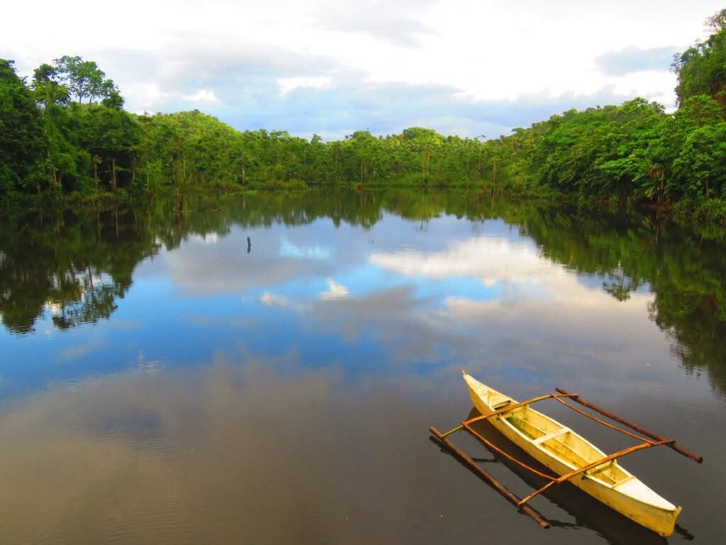 A tranquil lake surrounded by lush greenery reflects the sky, with a small wooden canoe with outriggers floating on the calm water.