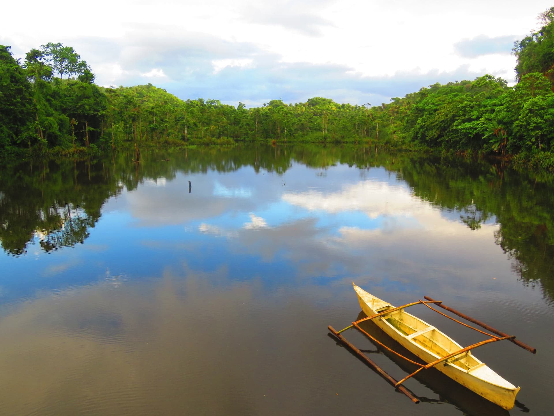 A tranquil lake surrounded by lush greenery reflects the sky, with a small wooden canoe with outriggers floating on the calm water.